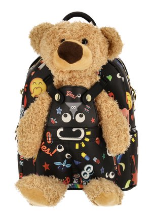 Ursuleț de pluș de culoare cămilă atașat de un rucsac negru cu modele colorate jucăușe de desene animate și emoji, inclusiv ochi, stele și pastile.