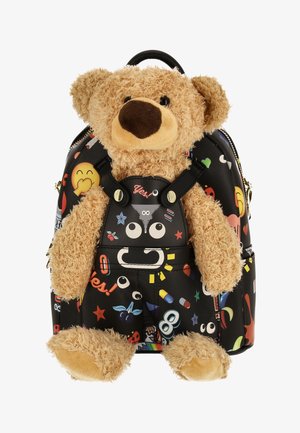 Beige teddybeer bevestigd aan een zwarte rugzak met kleurrijke, speelse cartoon- en emoji-patronen, waaronder ogen, sterren en pillen.