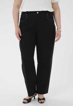 Person wearing black straight-leg pants with studded pockets, white top, black heeled sandals, a ring, and a silver bracelet on a white background.