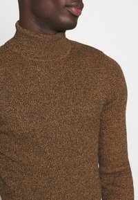 Gros plan d'une personne portant un pull à col roulé texturé marron, montrant le cou, l'épaule et une partie du visage de profil.