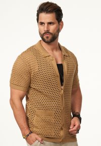 Beige gestrickte Strickjacke mit einem lockeren, offenen Webmuster, kurzen Ärmeln und einem Polokragen. Verfügt über zwei Fronttaschen und eine Knopfleiste.