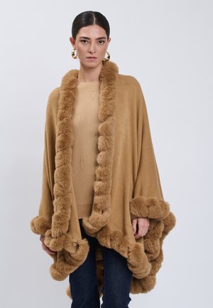 Frau mit dunklen Haaren, die goldene Creolen, einen braunen Pullover, blaue Jeans und einen braunen, mit Fell besetzten Umhang trägt und vor einem schlichten Hintergrund steht.