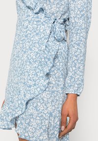 Närbild av en blå klänning med vitt blommönster, volanger, långa ärmar och en sidofrunt detalj vid midjan.