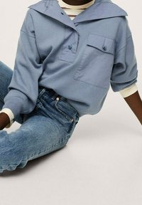 Persona che indossa una camicia azzurra con bottoni e tasca sopra una maglietta a righe e jeans blu, seduta con le gambe piegate su uno sfondo chiaro.