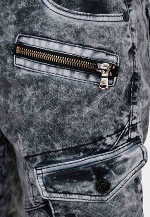 Pantaloni cargo in denim nero e grigio con una texture usurata. Le caratteristiche includono una tasca con zip metallica e dettagli di cuciture.