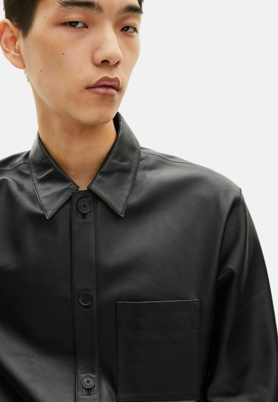 Camicia in pelle nera con colletto a punta, chiusura a bottoni e taschino a toppa. Texture liscia con spalle strutturate.