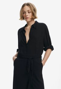 Chemise noire à boutons avec manches retroussées, fabriquée en tissu léger. Elle présente une ceinture à la taille, un décolleté en V profond et une texture verticale subtile.