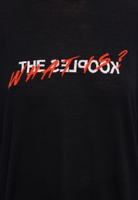 Czarny t-shirt z krótkim rękawem z wyraźnym białym i czerwonym napisem „WHAT IS? THE KOOKS” w stylizowanej czcionce. Miękka, gładka faktura.