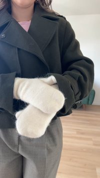 Manoplas de lana crema con una textura suave, sostenidas en la manga de un abrigo verde oscuro. Pantalones grises visibles abajo, suelo de madera en el fondo.