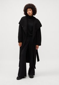 Cappotto nero sovradimensionato realizzato in un tessuto simile alla lana, con ampi rever e una vita con cintura, abbinato a un dolcevita nero e pantaloni a gamba larga.