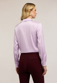 Kvinde med blondt hår iført guldsmykker, en langærmet lavendelfarvet bluse og højtaljede mørkerøde bukser, set fra siden.