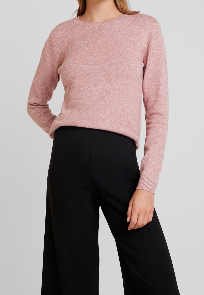 Femme portant un pull à manches longues rose pâle, rentré dans un pantalon noir taille haute à jambes larges, sur un fond neutre.