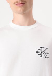 Balta medvilninė marškinėliai su apvalia iškirpte, su juodu siuvinėtu logotipu kairėje krūtinės pusėje, lygaus paviršiaus ir klasikinio silueto.