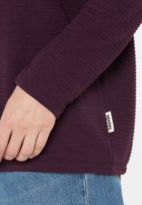 Lila strukturiertes Langarmoberteil mit einem taillierten Bund, das am unteren Saum ein kleines weißes Etikett mit der Aufschrift "SENSES" trägt.