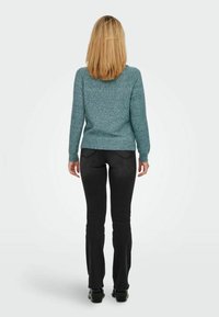 Türoffarbener Pullover mit langen Ärmeln und strukturiertem Strickmuster, kombiniert mit schwarzen Jeans im gerade geschnittenen Stil. Von hinten betrachtet.