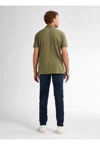 Olivově zelená polo tričko s krátkým rukávem, viditelný detail na zádech; kombinováno s tmavě modrými džíny a bílými teniskami. Minimalistický design po celém oblečení.
