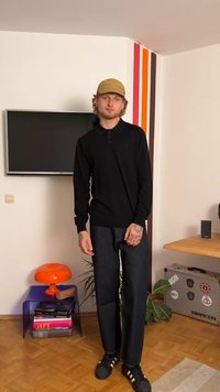 Schwarzes Langarm-Polohemd, dunkle Jeans, schwarze Turnschuhe; lockige Haare und eine beige Mütze tragend. Innenraum mit bunten Wandakzenten.