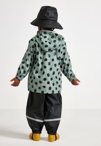 Vandtæt mintgrøn regnjakke med sorte prikker, parret med sorte regnbukser og gule støvler. Har en justerbar hætte.