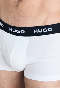 Weiße Boxershorts mit einem schwarzen Gummibund, der das weiße "HUGO"-Logo trägt. Glatte Textur und taillierte Passform, geschnitten über dem Oberschenkel.