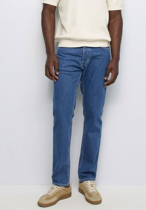 Homme portant un jean droit bleu, des baskets beige et marron, et un t-shirt tricoté blanc à manches courtes, debout contre un mur blanc uni.