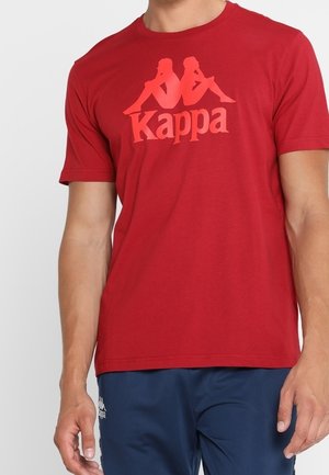 Mężczyzna noszący czerwoną koszulkę Kappa z logo dwóch siedzących plecami do siebie postaci oraz granatowe spodenki.