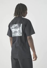 Mann mit Dreadlocks steht seitlich und trägt ein schwarzes T-Shirt mit einem schwarz-weißen Grafik von einem Truck und Fahrradreifen auf dem Rücken.