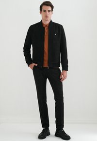 Sort softshell-jakke med lynlås, båret over en rustfarvet polo. Sættet inkluderer teksturerede mørke bukser og sorte sneakers.