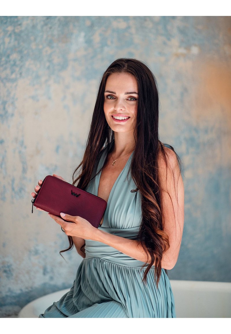 Burgundy leather wallet with a zip closure, featuring a black embossed logo. Set against a textured blue-green background.