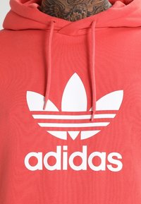 Felpa con cappuccio corallo con logo Adidas bianco, caratterizzato da tre foglie e strisce orizzontali. Realizzata in un tessuto morbido, con cappuccio regolabile.