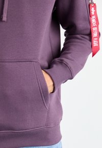 Sudadera morada con bolsillo tipo canguro, puños y dobladillo acanalados, con una etiqueta de tela roja que dice "QUITAR ANTES DEL VUELO." Textura suave.