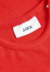 Rotgeripptes Gewebe mit rundem Halsausschnitt. Das Etikett zeigt den Text "JJXX" in Marineblau auf weißem Hintergrund, positioned innerhalb des Kragens.