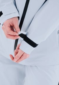 Ljusblå vattentät jacka med svart kardborrejustering vid ärmarna. Slät yta och figursydd design. Tydlig logotyp på ärmen.