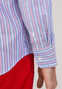 Camisa de rayas azul y blanca con acentos rojos, que cuenta con un puño abotonado y un dobladillo recto. La tela parece texturada y ligera.