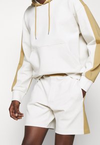 Vit hoodie med guldakcenter, med en framficka och snoddar. Matchas med vita shorts. Mjuk, slät tyg.