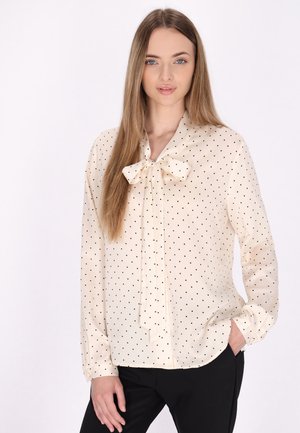 Blusa crema con pois neri, presenta un collo a fiocco, maniche lunghe, orlo curvo e tessuto leggero.