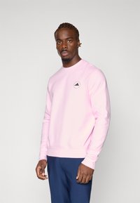 Sweatshirt rose en tissu doux, avec un col rond, des manches longues et un petit logo noir sur la poitrine.