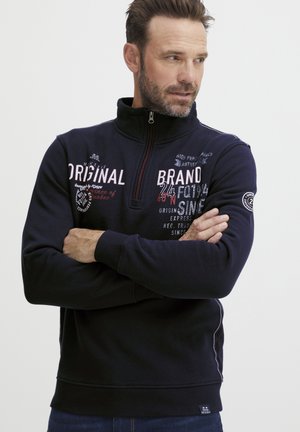 Sudadera con cremallera de color azul marino y cuello alto, con diseños gráficos de texto en blanco y rojo y un parche de logo redondo en la manga izquierda.