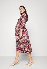 Vestido midi em padrão de leopardo nas tonalidades rosa, preto e dourado, com mangas compridas e cintura ajustada, combinado com sandálias de salto amarelo pálido.
