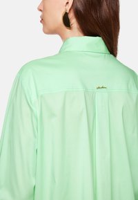 Mintgrünes Shirt mit lässiger Passform, das einen spitzen Kragen, ein Schulterdetail und ein kleines goldenes Logo auf der Rückseite aufweist.