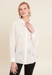 White button-up shirt featuring embroidered cut-out floral patterns, long sleeves, and a rounded collar. Black pants are worn beneath.