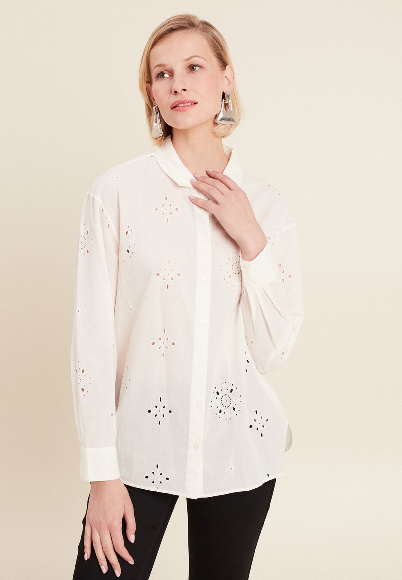 White button-up shirt featuring embroidered cut-out floral patterns, long sleeves, and a rounded collar. Black pants are worn beneath.