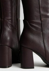 Stivaletti in pelle marrone con una texture liscia, tacco a blocco, cerniere laterali e dettagli di design curvi sulla gamba.