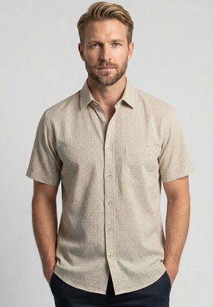 Man met baard die een beige overhemd met korte mouwen en subtiel patroon draagt, staand met handen in de zakken tegen een effen achtergrond.