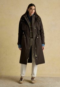 Hnedý dlhý kabát s nastaviteľným pásom, viacerými prednými vreckami, zipsom a záklopkami. Prehodený cez vzorovaný sveter a košeľu. Svetlé nohavice a tenisky.