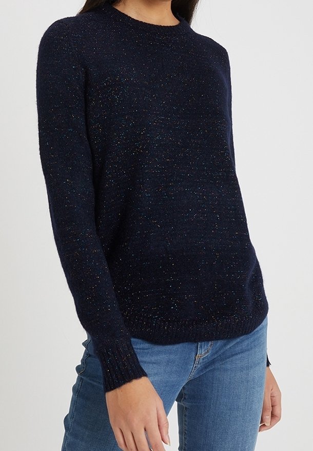 Femme portant un pull tricoté bleu foncé à motifs tachetés avec manches longues, associé à un jean bleu clair sur un fond uni.