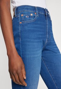 Vaqueros de mezclilla azul con corte slim, que cuentan con herrajes de cobre, un parche de logo característico y un leve desgaste a lo largo de las piernas.