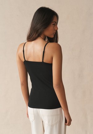 Femme aux longs cheveux bruns portant un top noir à fines bretelles et un pantalon blanc, de profil sur un fond beige uni.