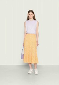 Top sem mangas lavanda combinado com uma saia midi em camadas amarela com flores. Modelo a usar tênis brancos e a segurar uma pequena bolsa roxa.