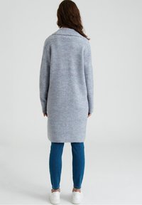 Cardigan lungo grigio con un motivo di maglia texturizzato e un ampio colletto, abbinato a jeans skinny blu e sneakers bianche.
