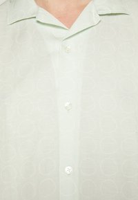 Camicia verde chiaro con bottoni, realizzata in tessuto strutturato con un sottile motivo circolare, sei bottoni bianchi e un design con colletto classico.